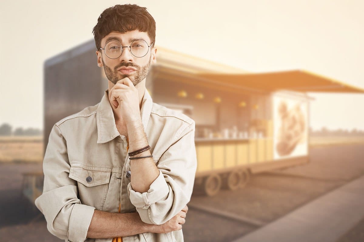 Young man business owner in glasses and jacket standing in front of a food cart parked in a parking lot