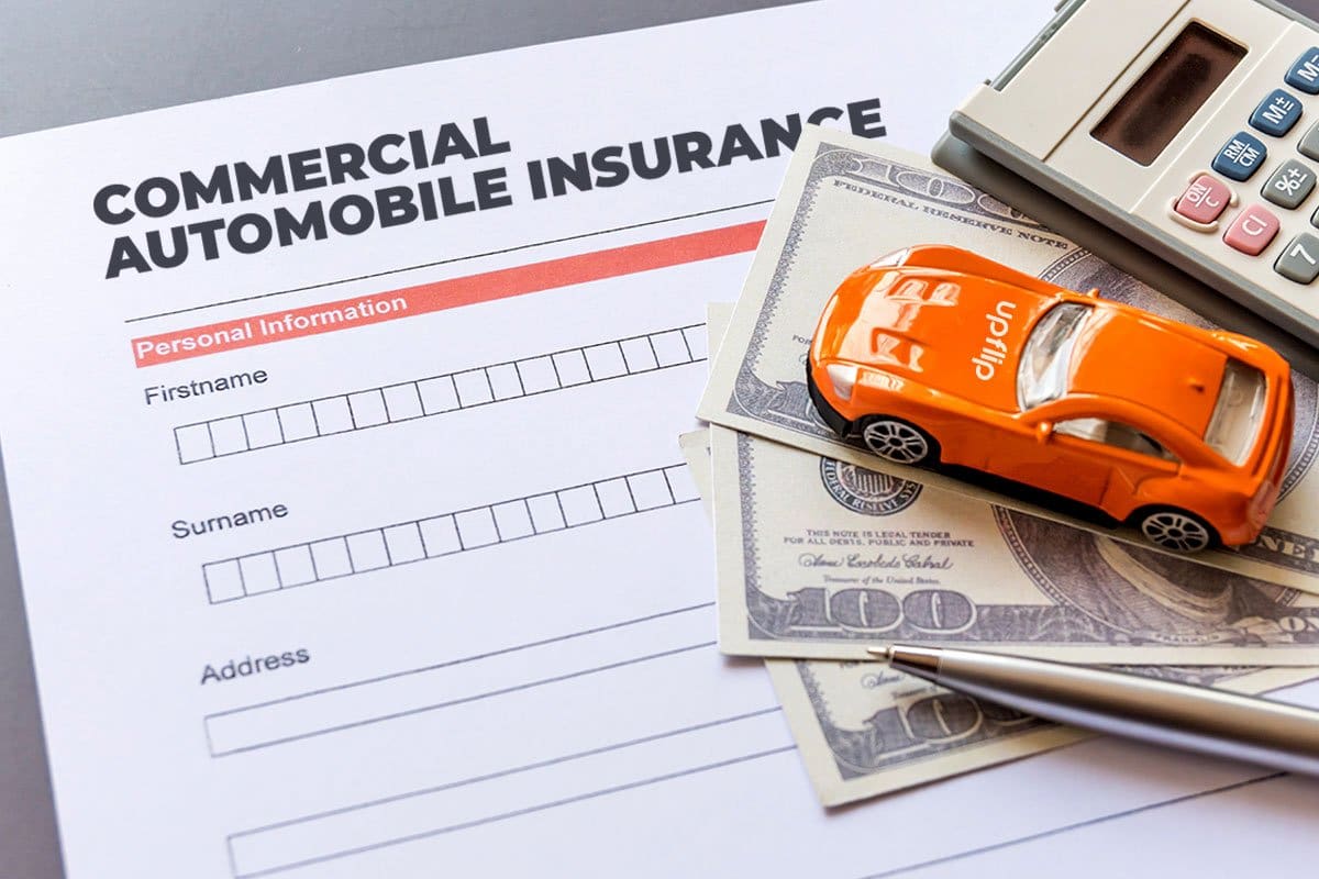 Lay-flat shot of a commercial automobile insurance form on a table with hundred dollar bills, a toy car, a pen, and a calculator on top