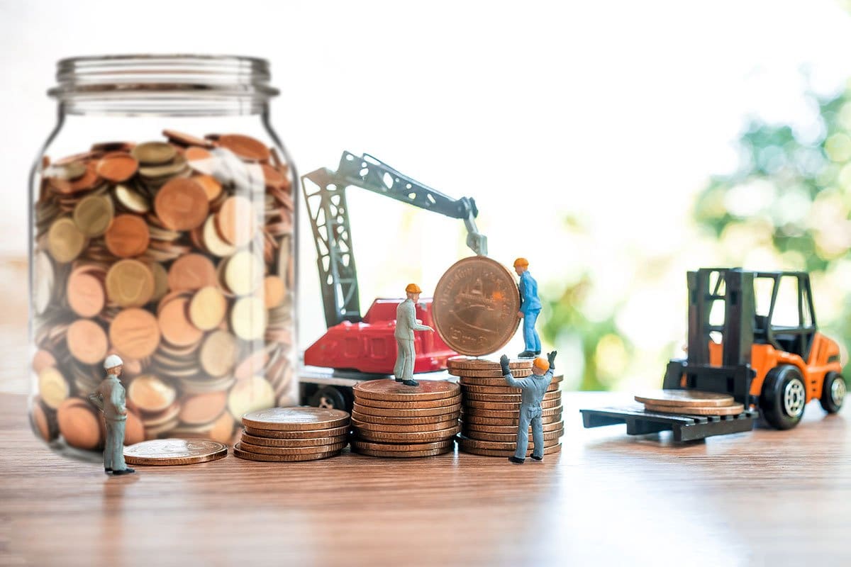 Financial institutions concept showing miniature toy construction workers using a crane and forklift to move stacks of coins next to a full jar of coins