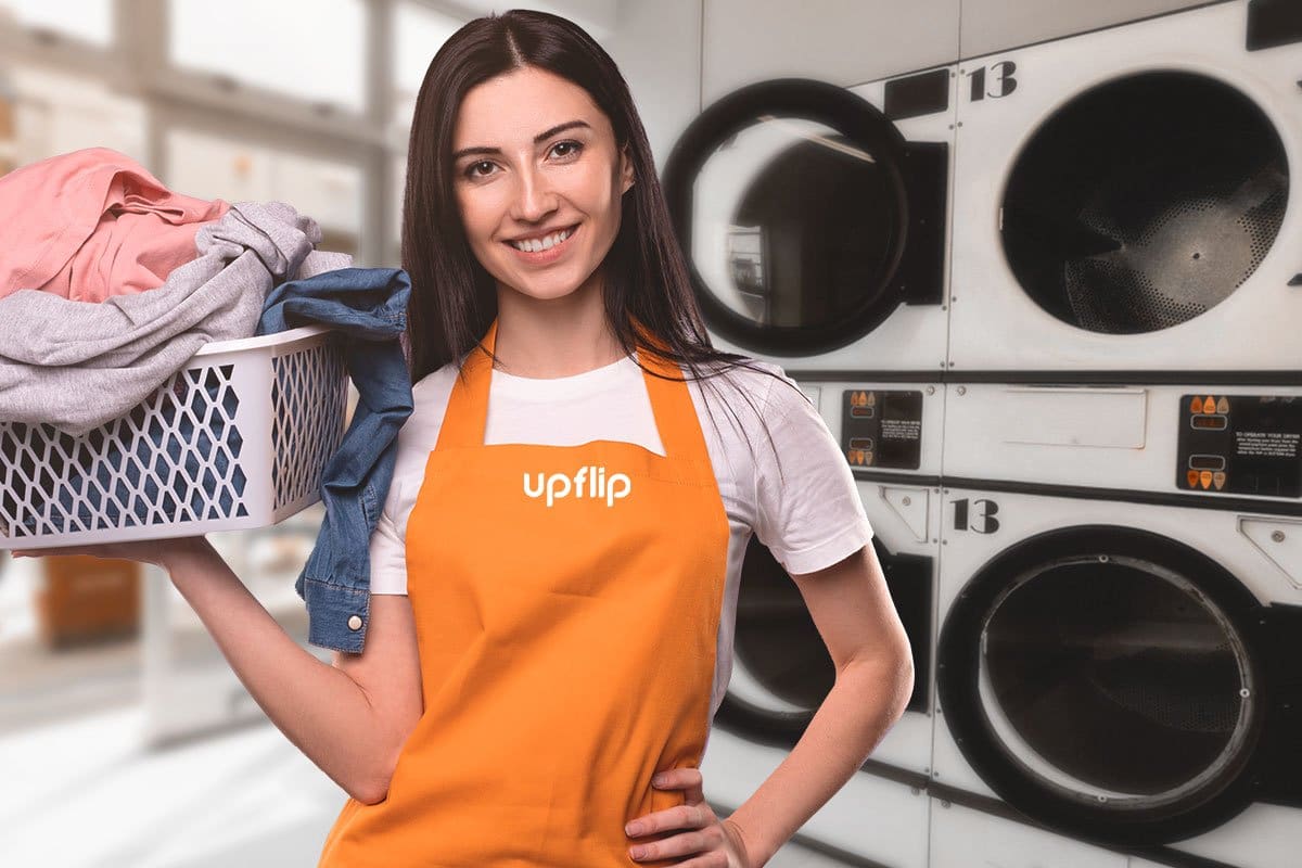 Laundromat attendant in an orange apron standing in front of washing machines and holding a basket of freshly dried clothing