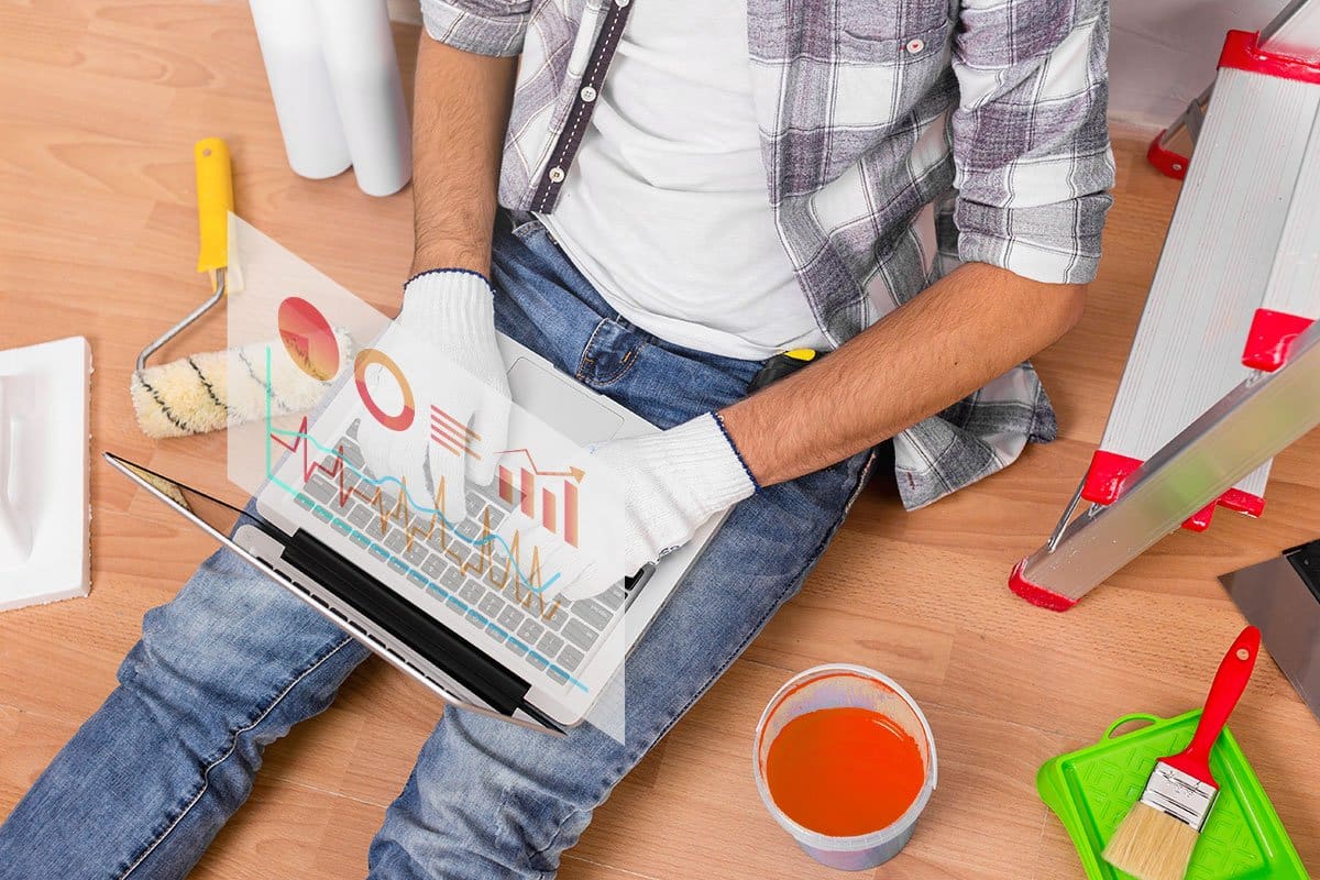 Top-down shot of a painter sitting on the floor surrounded by painting gear and a ladder doing quick research on a laptop