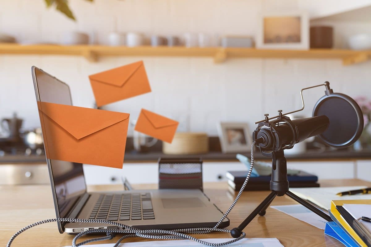 How to Grow a Podcast (2025) 11 Podcast email marketing concept showing orange envelopes flying out of a laptop on a table with a professional microphone, notepads, and binders