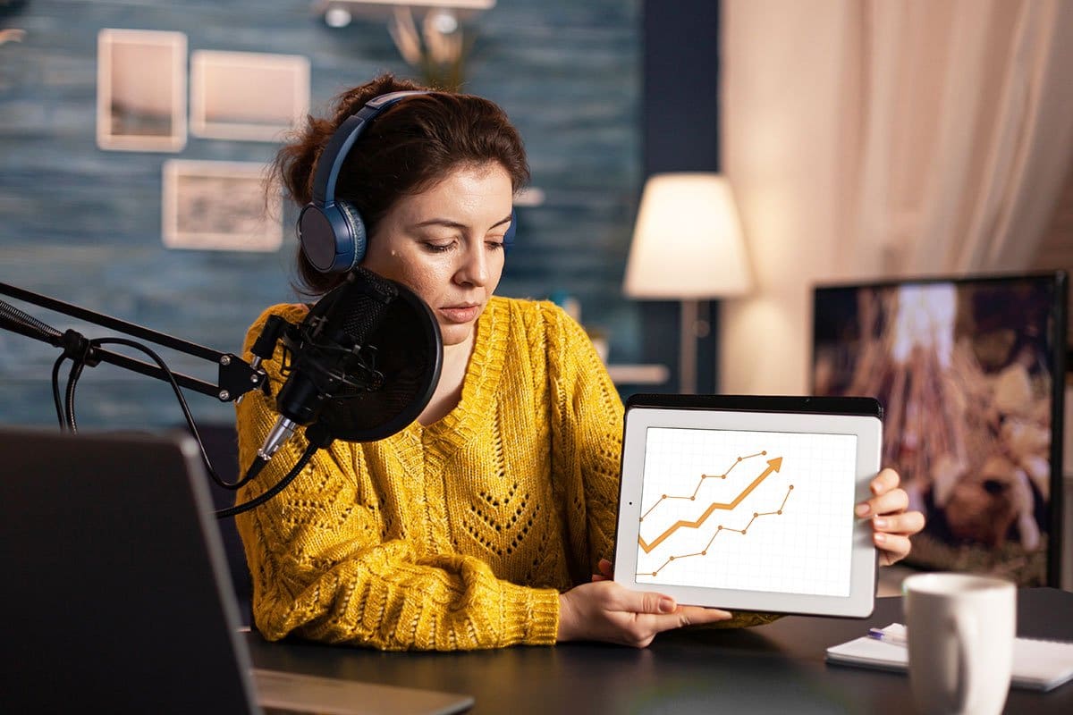 How to Grow a Podcast (2025) 15 Woman podcaster holding a tablet with an upward trending graph on it while sitting at her recording station