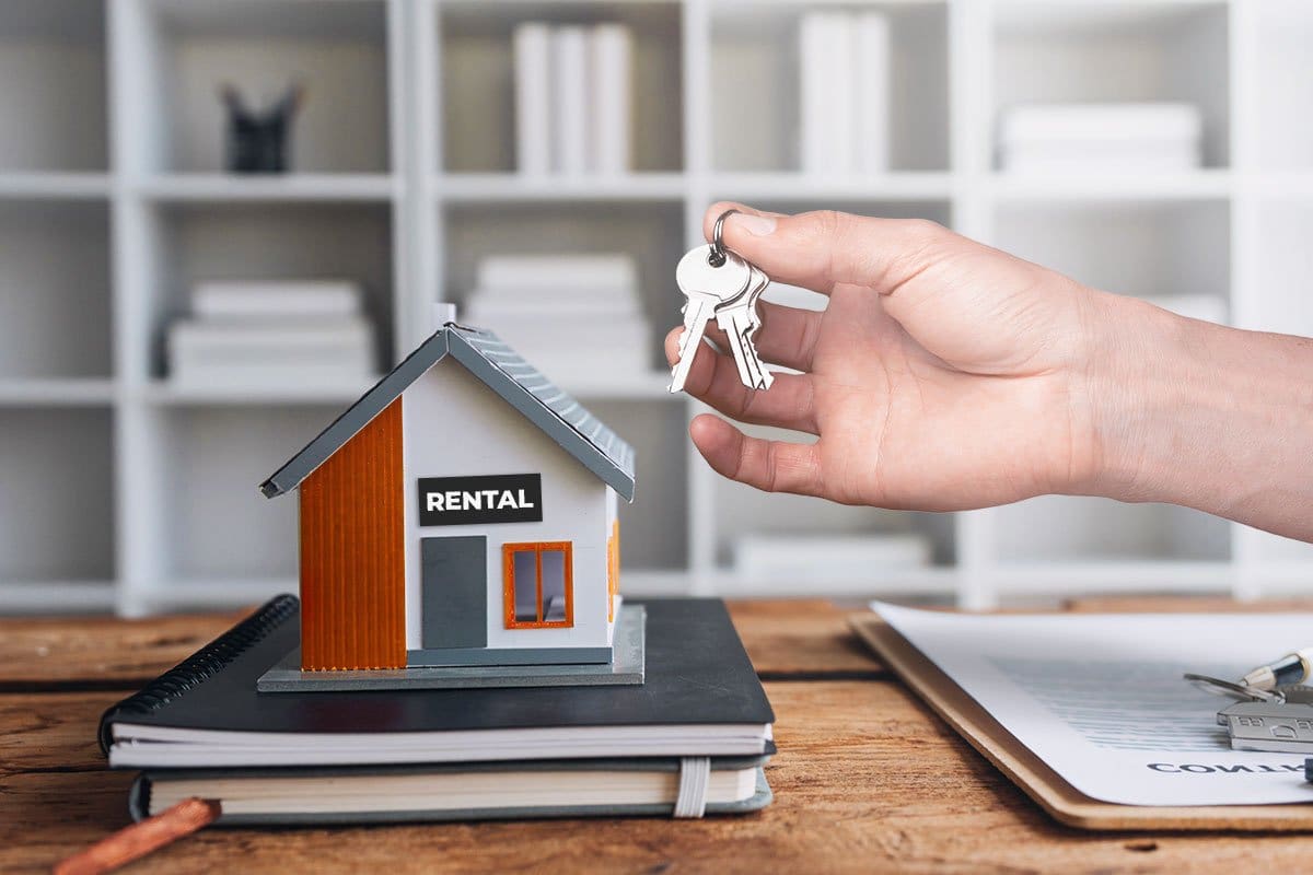Hand holding keys over a desk with a stack of notepads and a miniature house