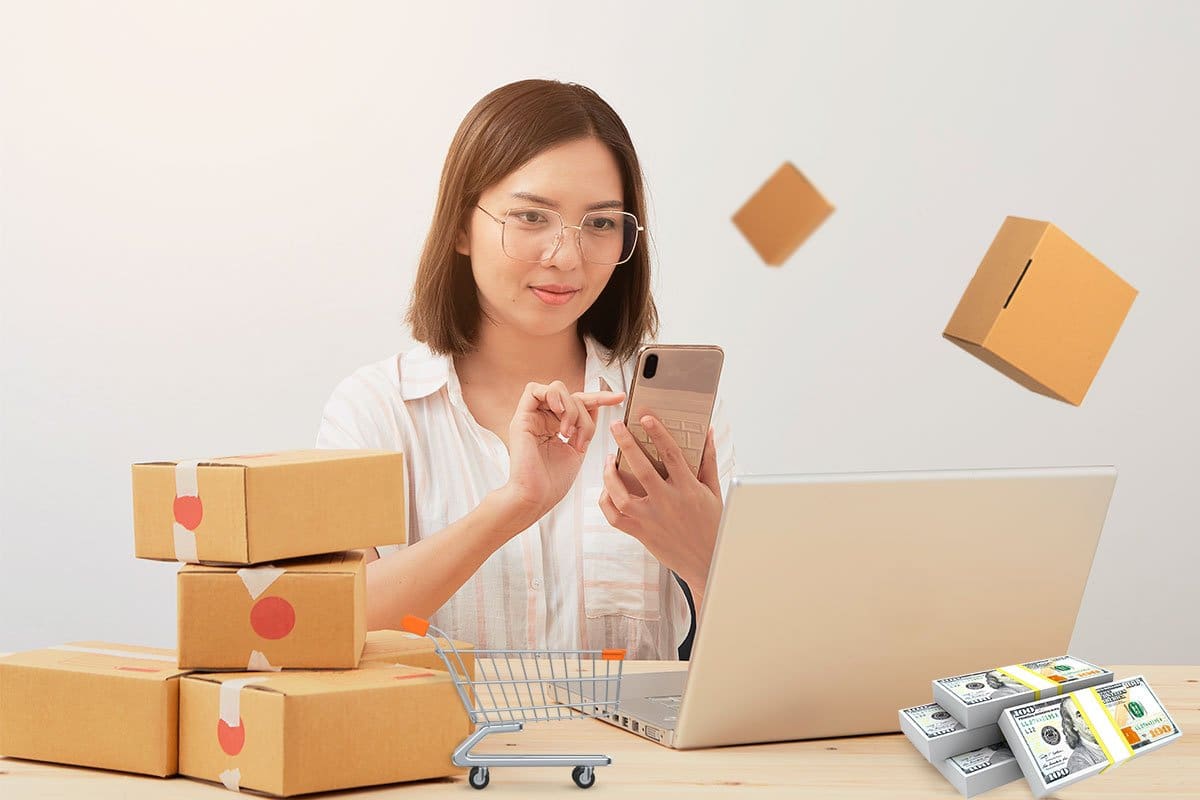 Woman using a smart phone while sitting at a desk with a laptop, boxes ready for shipping, stacks of cash, and a miniature shopping cart