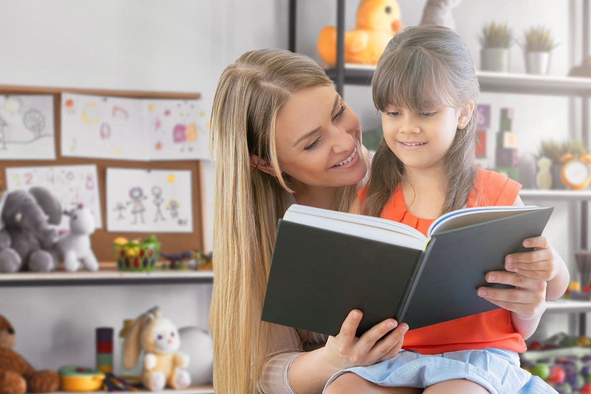 Young woman daycare owner reading a book to a preschool-aged girl