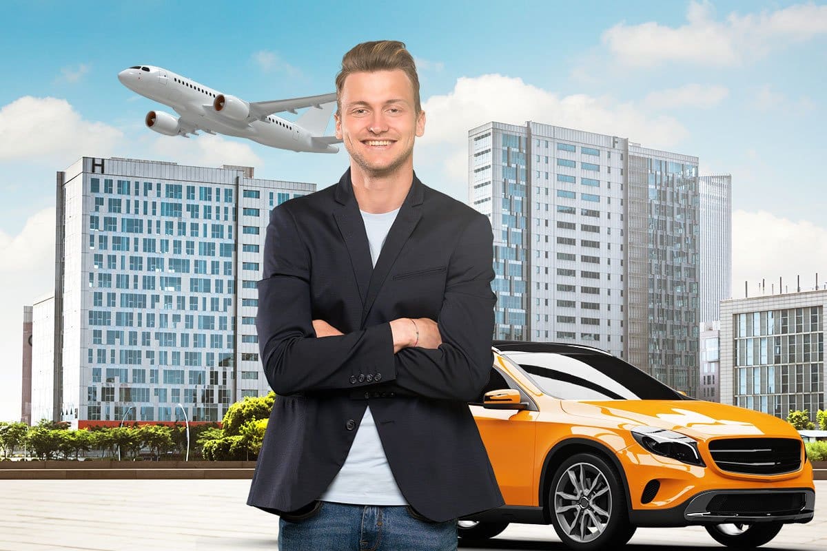 Businessman in a sports coat and jeans smiling in front of a rental car with a cityscape and a jet plane in the sky in the background