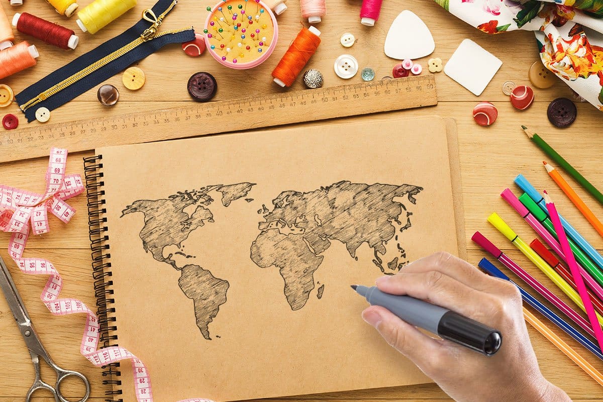 437 Stand-Out Craft Business Names 4 Top-down shot of an artist sketching a world map in a craft paper sketch book surrounded by colored pens, a wood ruler, scissors, a zipper, buttons, and pins