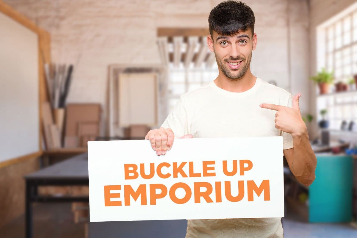 522 Etsy Shop Names You’ll Love (2025) 5 A man in a workspace holding a sign that reads "Buckle Up Emporium"
