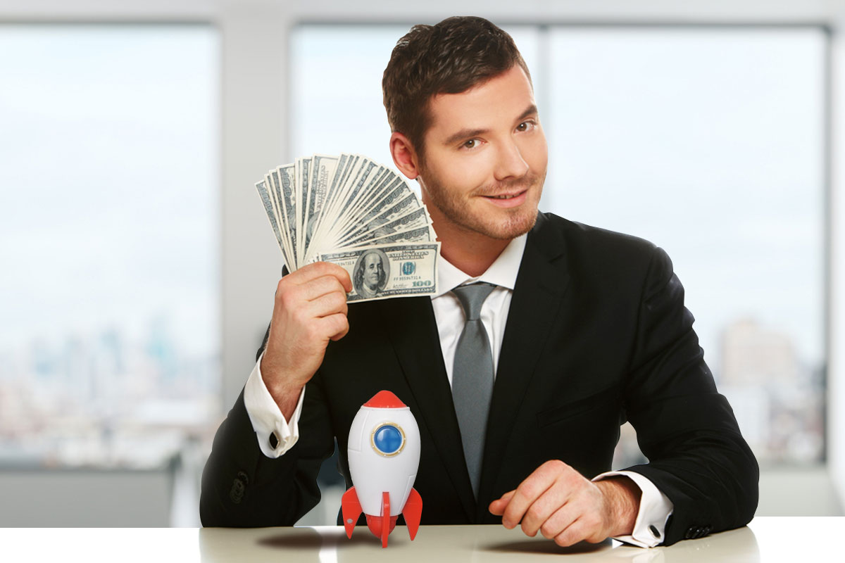 Sharply dressed business man holding a fan of cash in his hand with a miniature rocket ship sitting on the table in front of him.