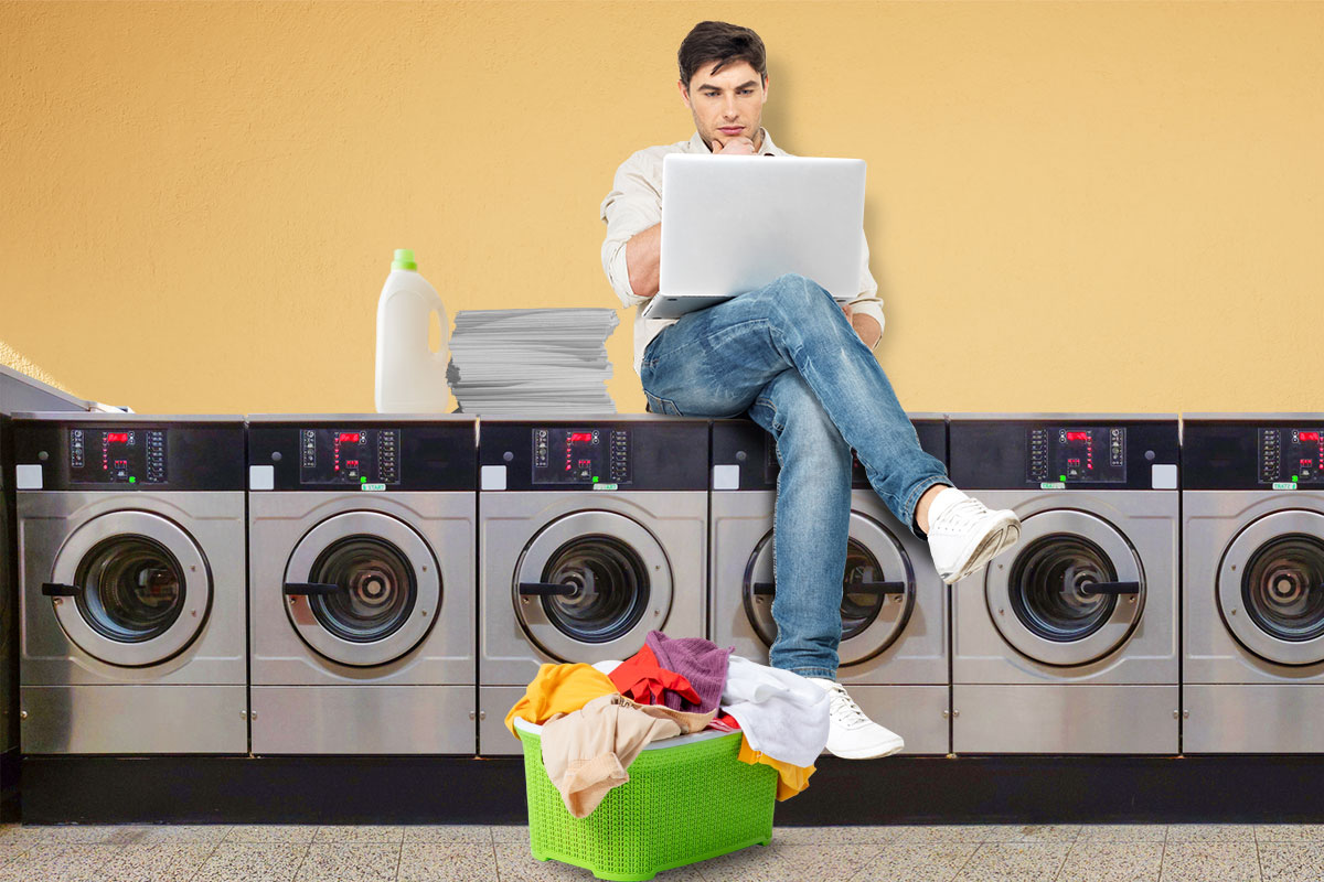 Man sitting on top of a small washing machine in a laundromat using a laptop