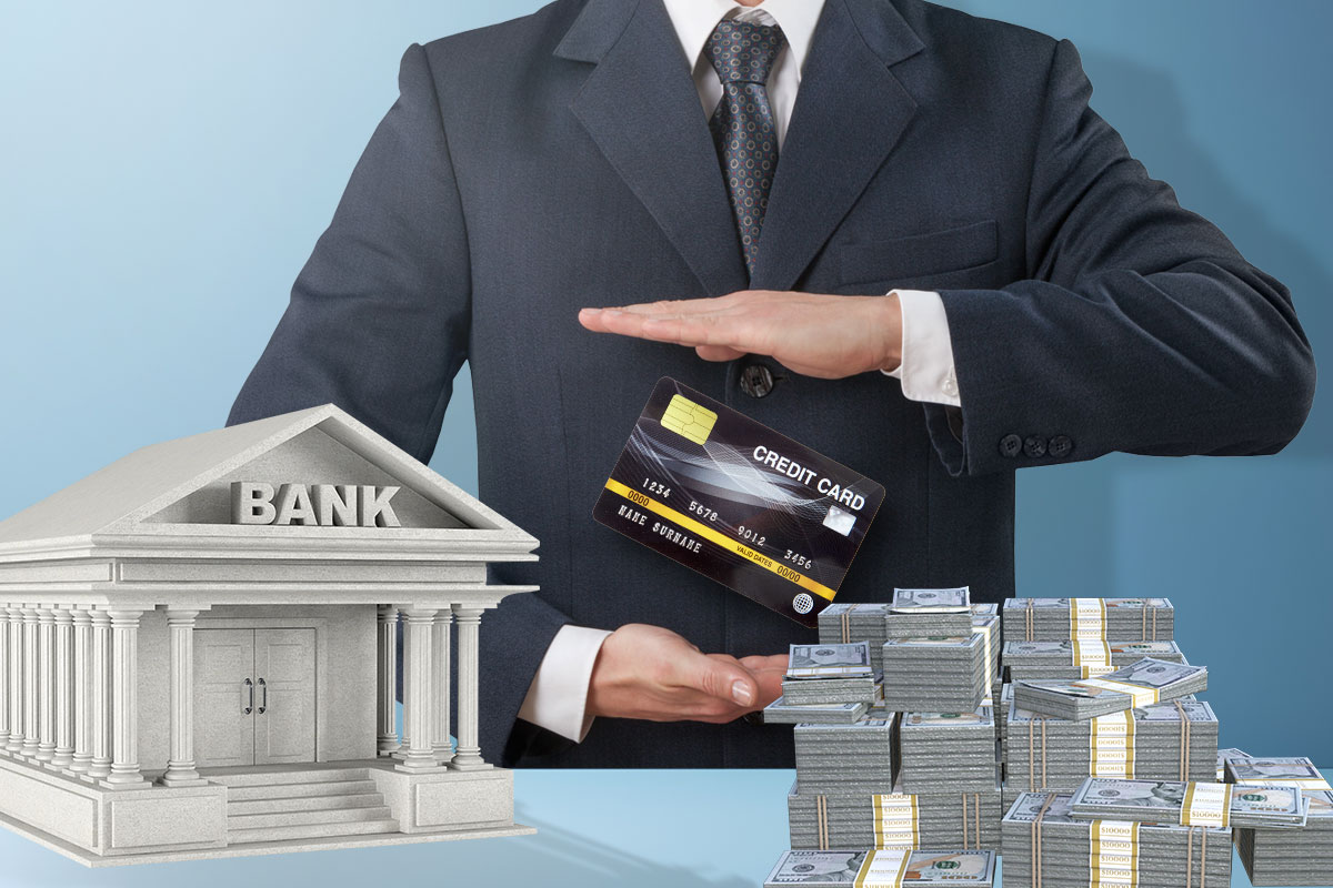Businessman in a suit with a credit card hovering between his hand standing behind a table with a miniature bank model and stacks of cash