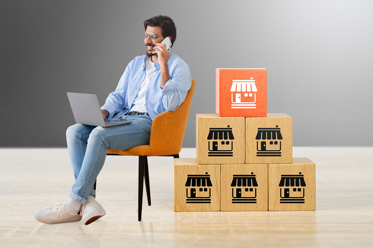 Turnkey Business: The Ultimate Guide (2025) 3 Casually dressed man working on a laptop while taking a call next to a stack of oversized wooden blocks with storefronts printed on them