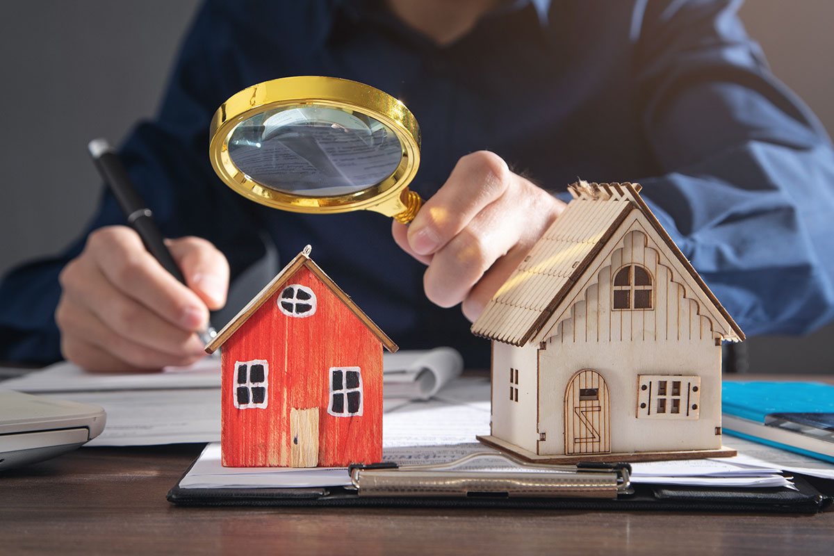 Business person taking notes while holding a magnifying glass over two miniature houses