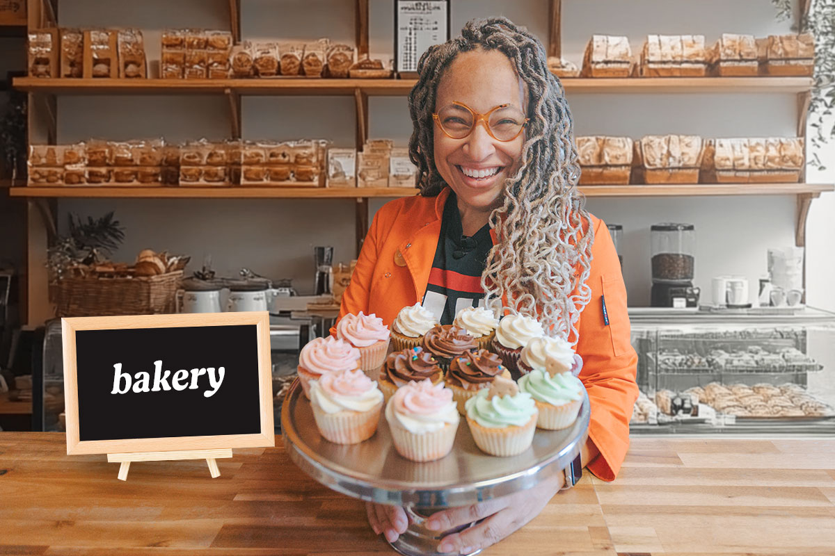 Owner of The Cupcake Collection standing at a counter holding a tray of cupcakes next to a sign with the word "bakery"