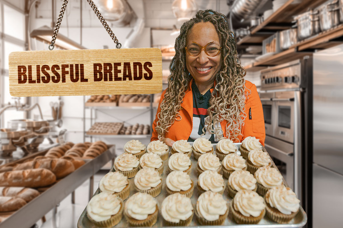 A woman in a bakery kitchen holding a tray of cupcakes next to a sign that says "Blissful Breads"