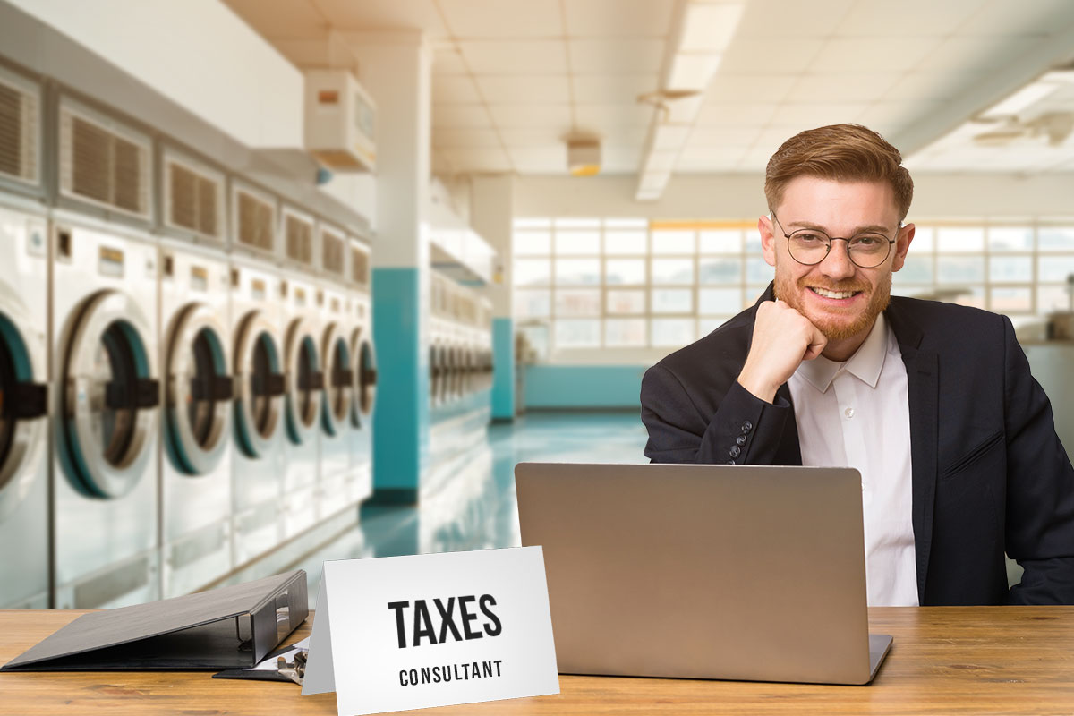 A businessman in a suit working at a laptop in a laundromat with a sign that reads "taxes consultant"