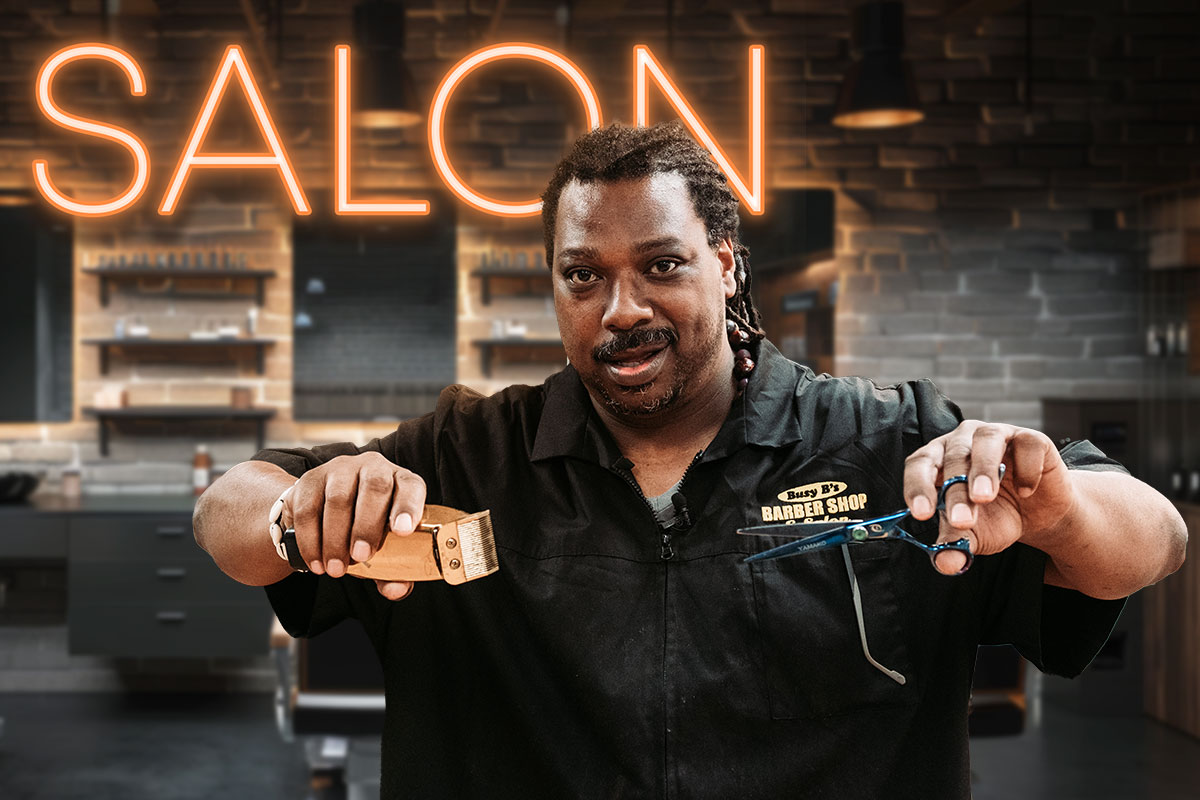 The owner of Busy B's Barbershop holding clippers and shears in front of a neon sign with the word salon.
