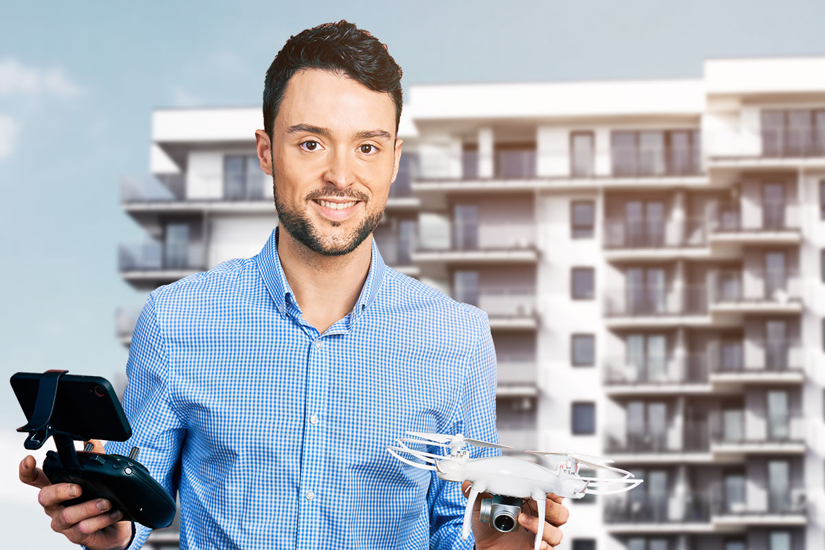A realtor holding a drone and a controller in front of an apartment complex.