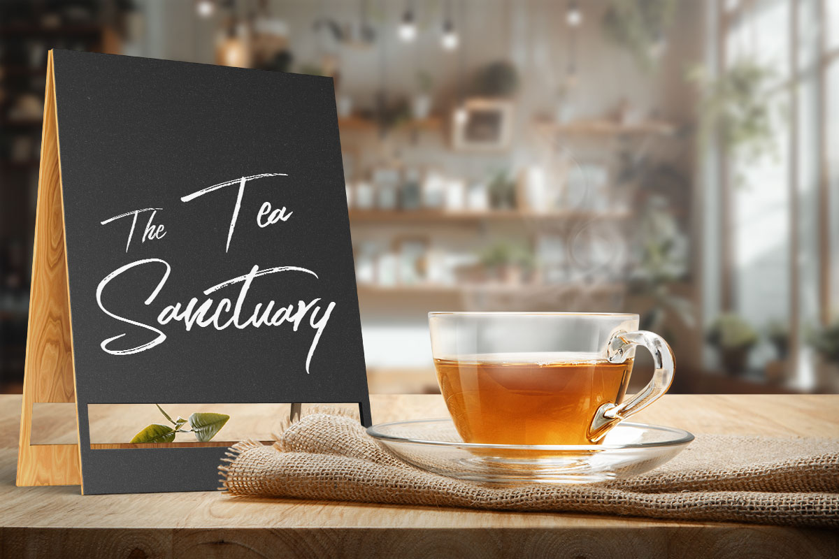 379 Coffee Shop Names You’ll Adore! 6 A cup of tea sitting on a counter in a tea shop next to a wooden sign that says The Tea Sanctuary.