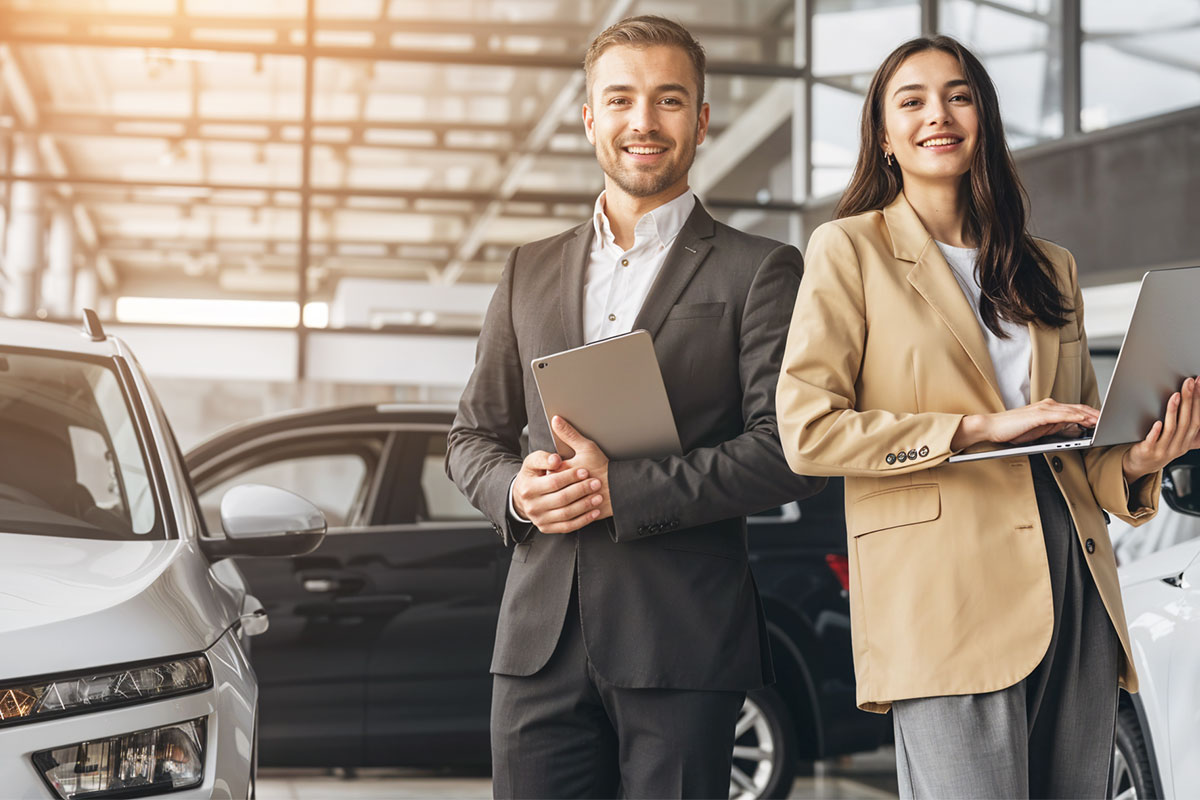 How to Start a Used Car Dealership 15 Two dealership employees standing next to cars while holding a tablet and a laptop.