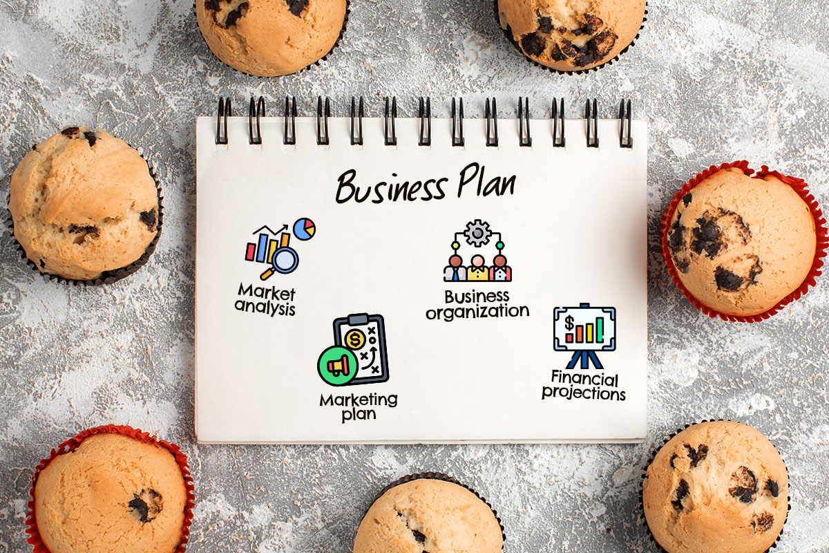 A notebook showing the stages of a business plan with cartoon drawings sitting on a counter next to baked goods.