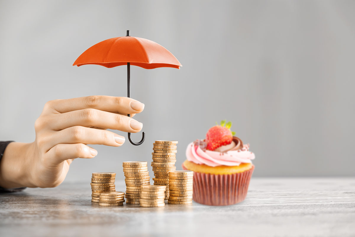 A hand holding a mini umbrella over a cupcake and stacks of coins.