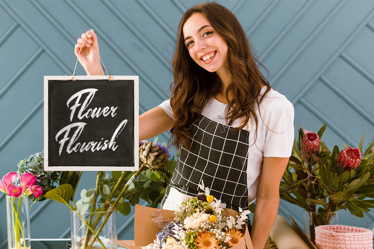 A flower shop owner surrounded by flowers holding a sign that says Flower Flourish.