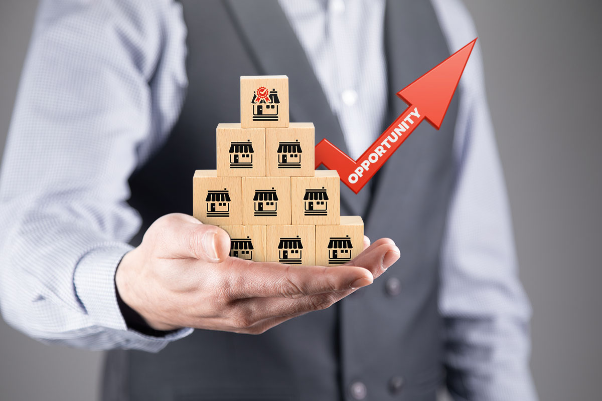 A business owner holding a stack of blocks with store symbols next to an upward arrow with the word opportunity on it.