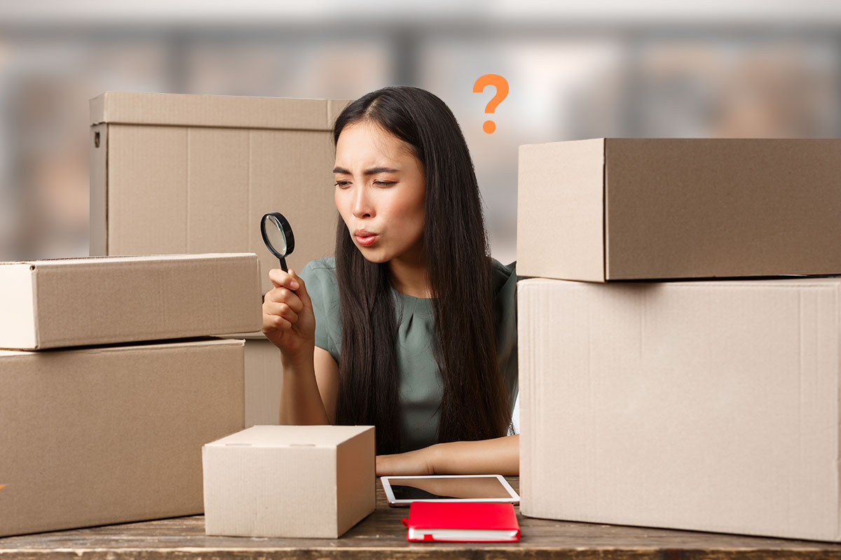 A woman with a magnifying glass analyzing a cardboard boxes piled up around her.