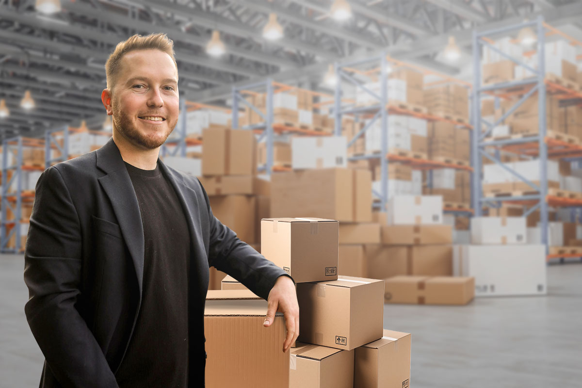 A dropshipping business owner leaning on a stack of boxes in a large warehouse full of products.