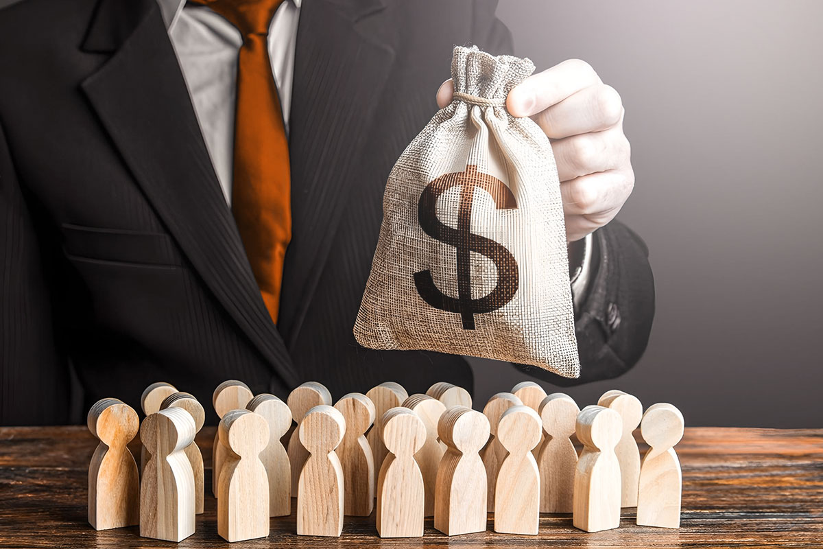 Grants 101 for Small Business Owners 74 A businessman holding a bag of money next to a desk with a group of people-shaped wooden cutouts.