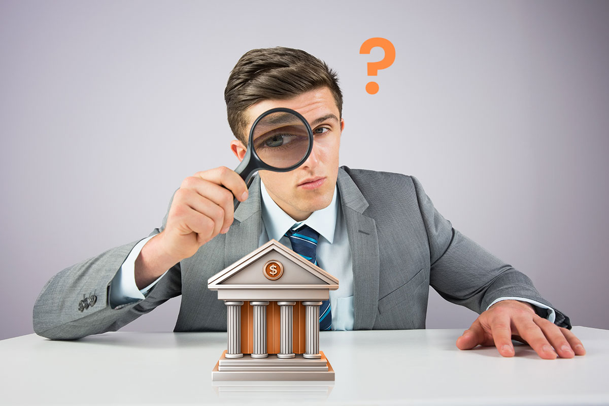 A businessman using a magnifying glass to inspect a model bank while a question mark hovers overhead.