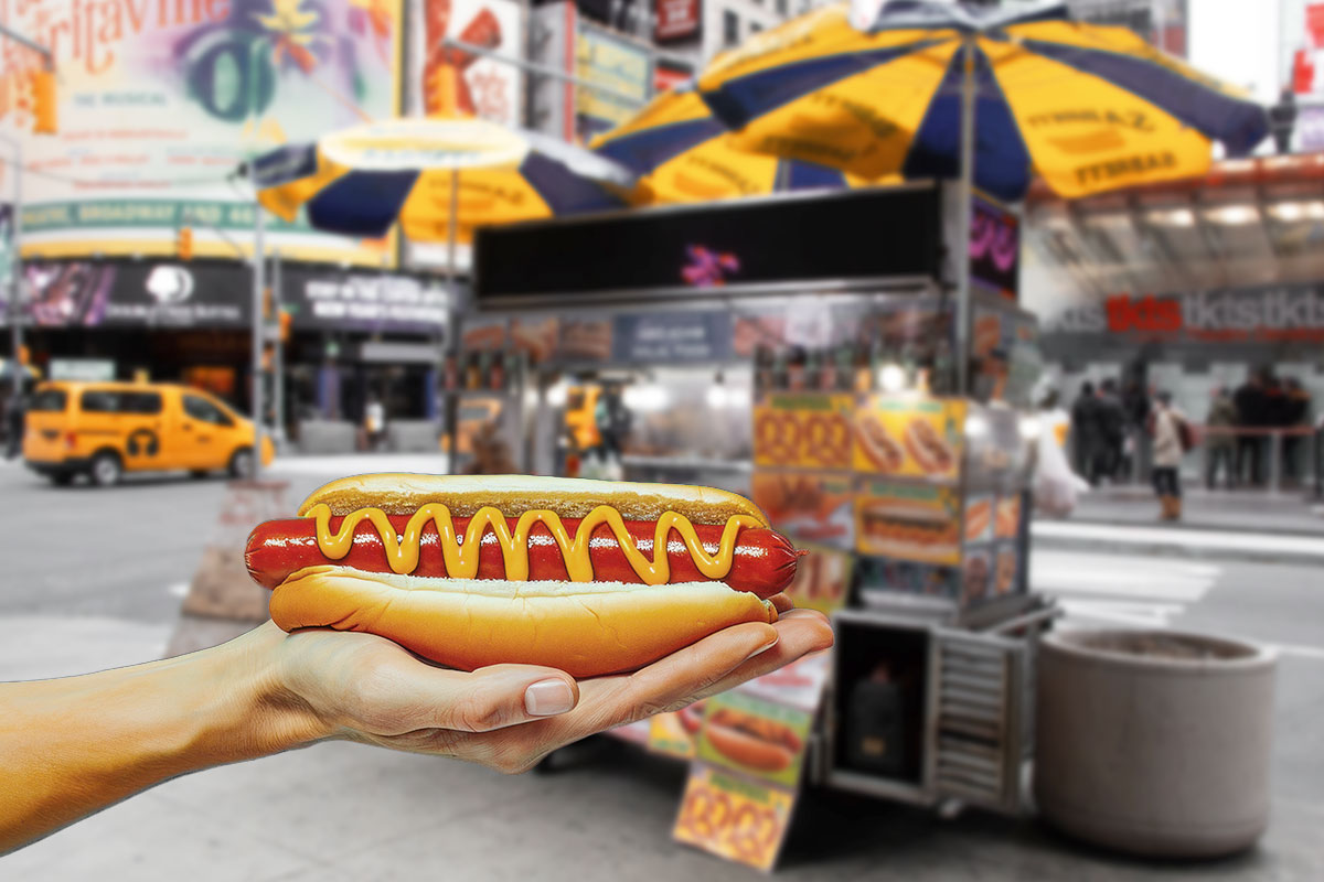 A person holding a fresh hot dog with mustard while standing in front of a hot dog cart.