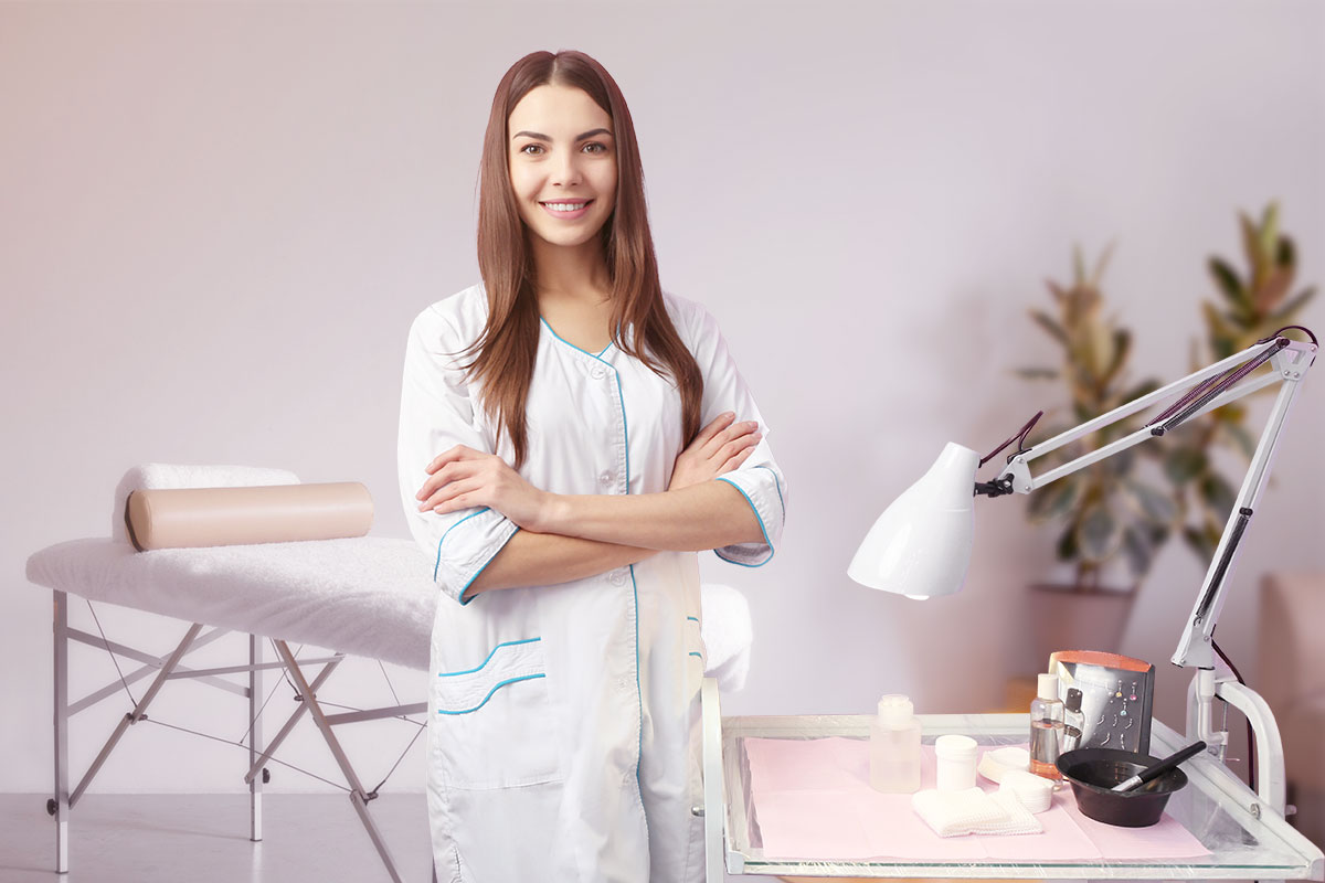 387 Beautiful Esthetician Business Names 2 An esthetician in a white gown standing next to a cart with a lamp and beauty supplies.