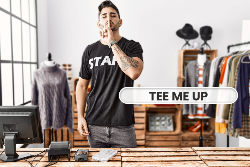 A businessman in a T-shirt holding a finger vertically across his lips next to a bubble that says Tee Me Up.